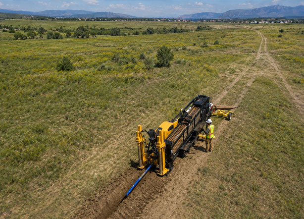 Drone aerial view of horizontal directional drilling rig installing underground pipeline across open rural land with construction worker overseeing trenchless utility boring operations.